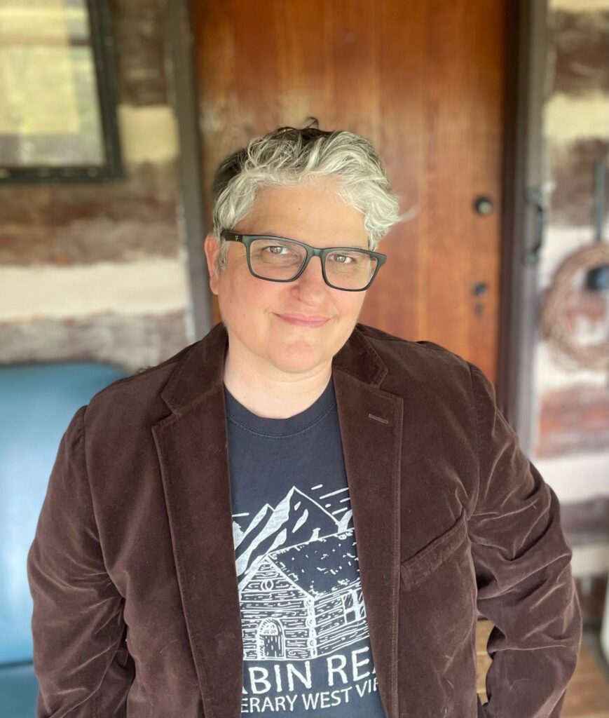 Image of children's book author Vicki Johnson in front of the front door of a log cabin.